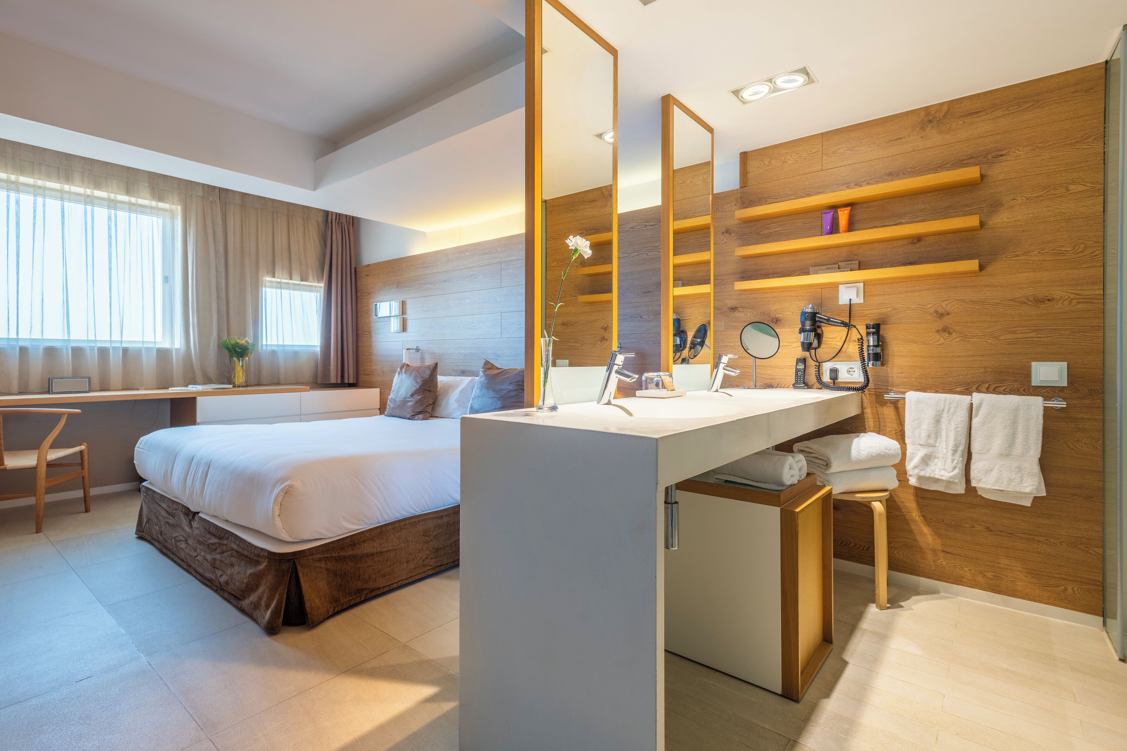 A modern hotel room with a large bed, a desk, a vanity with a mirror, and a wooden wall.