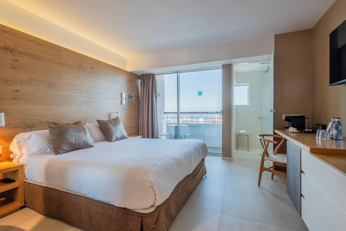 A modern hotel room with a large bed, a desk, and a balcony with a view.