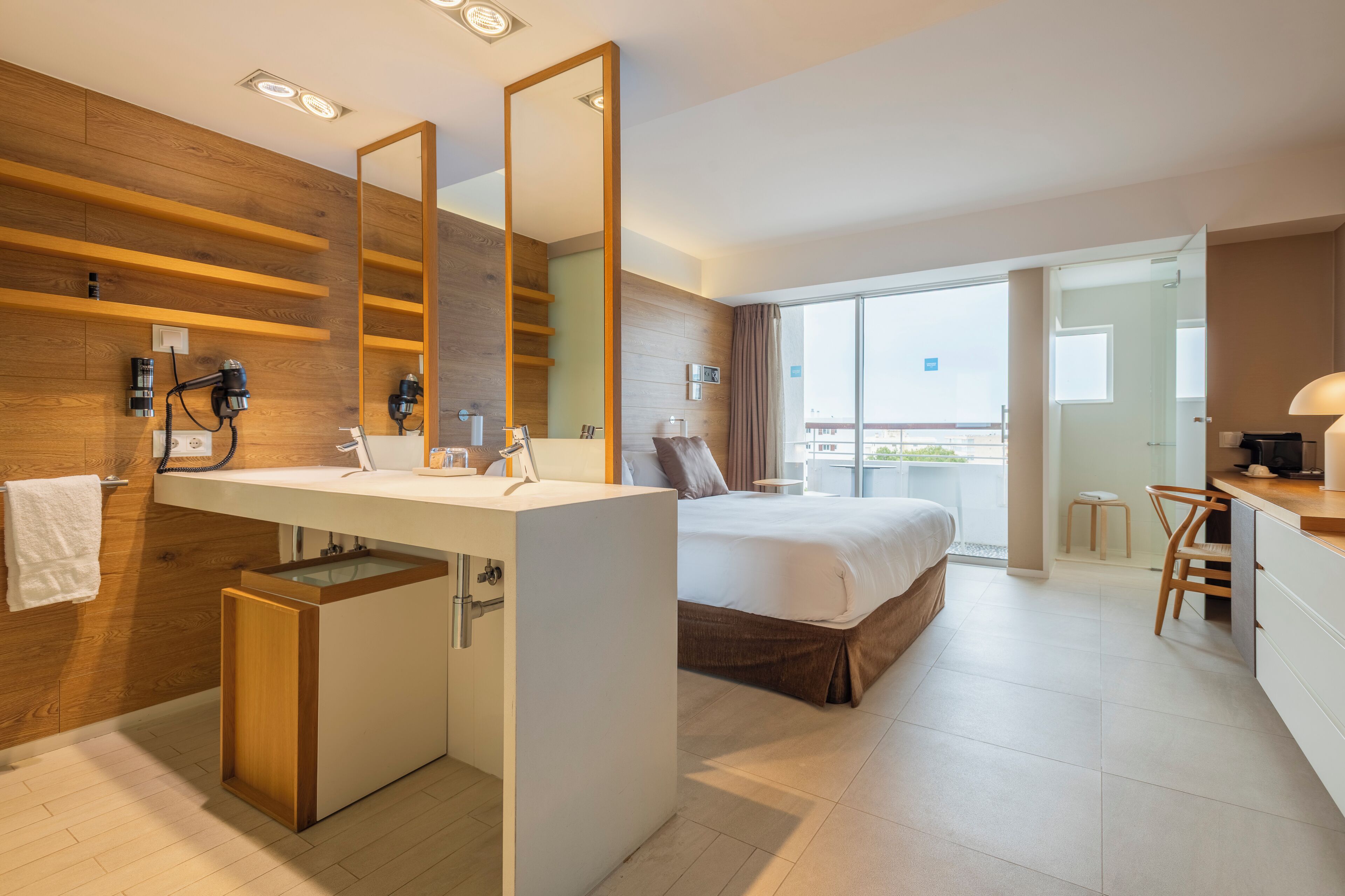 A modern hotel room with a wooden accent wall, a large bed, a separate bathroom with a vanity, and a balcony view.