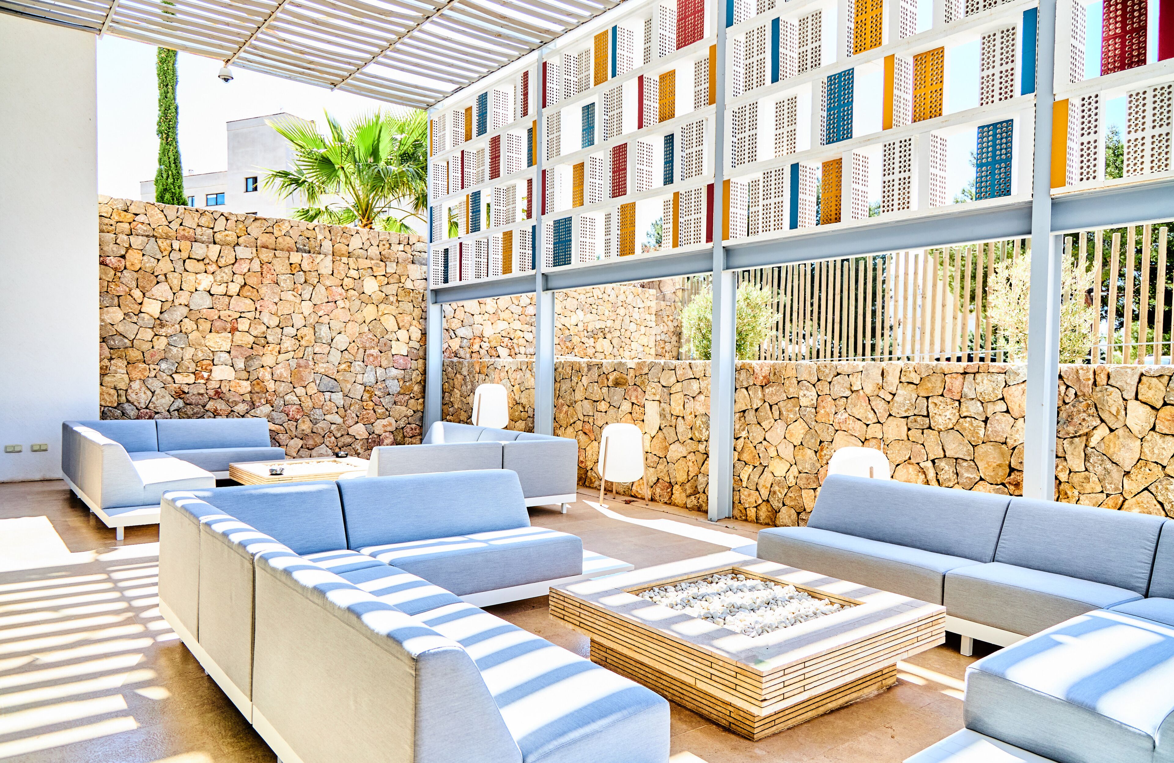 A modern outdoor lounge area with stone walls, blue cushioned seating, and a large window with colorful lattice panels.