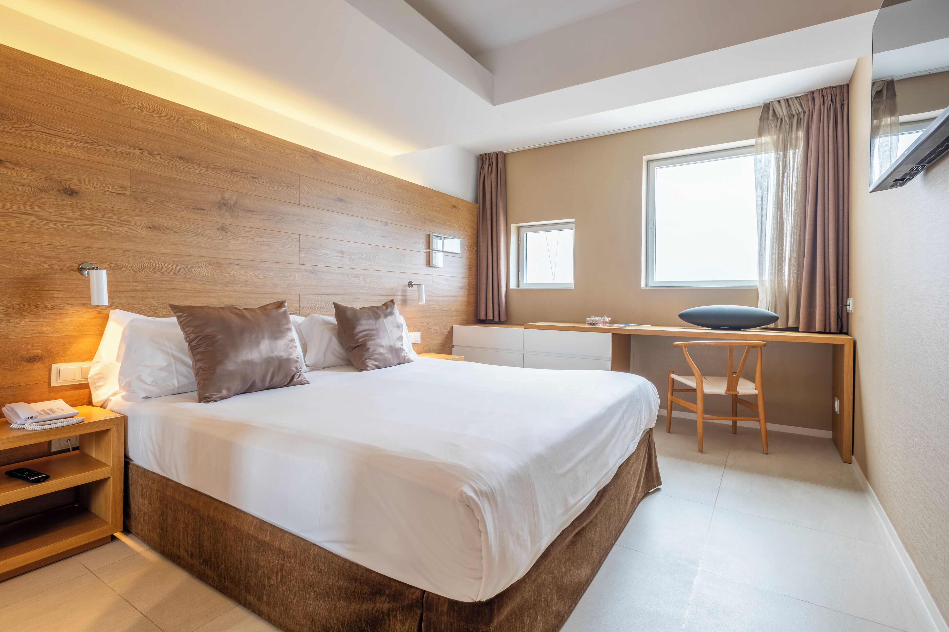 A modern hotel room with a large bed, wooden headboard, bedside table, and a desk with a chair.