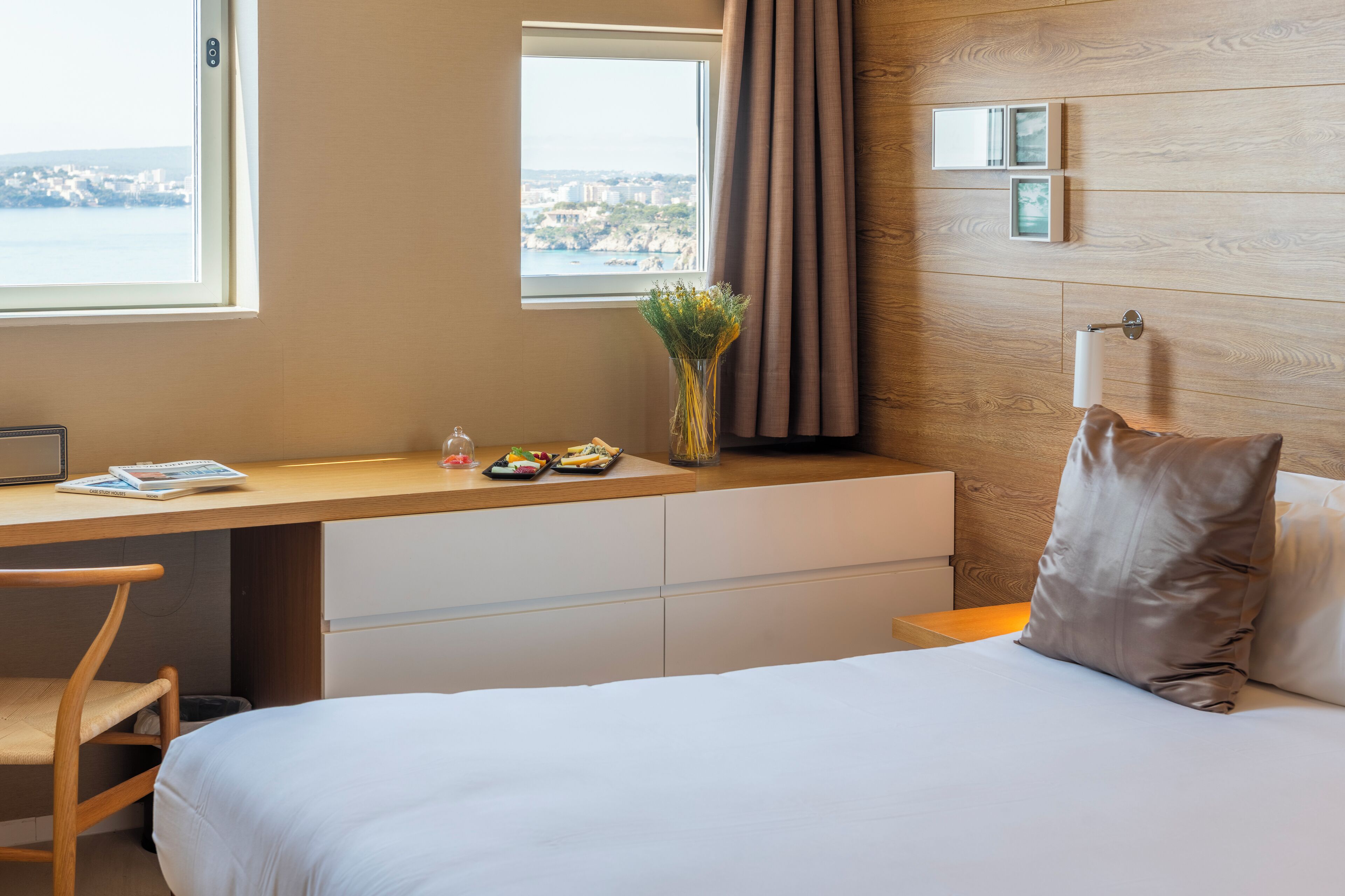 Quarto de hotel com cama, escrivaninha, cadeira, janela com vista, vaso de flores e mesa de cabeceira.