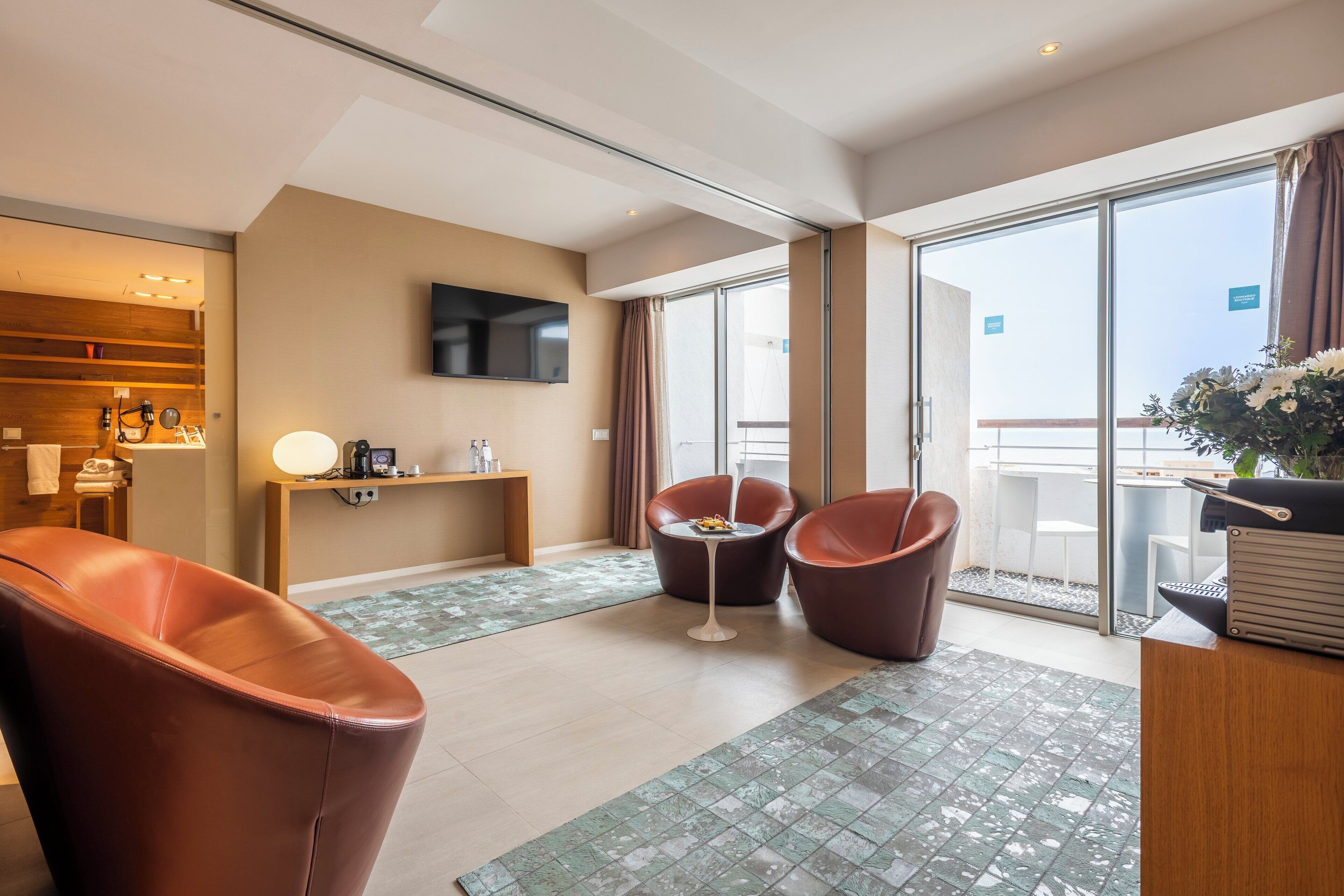 Una habitación de hotel moderna con un amplio ventanal corredero que da a un balcón, un televisor de pantalla plana en la pared y dos sillones de cuero.