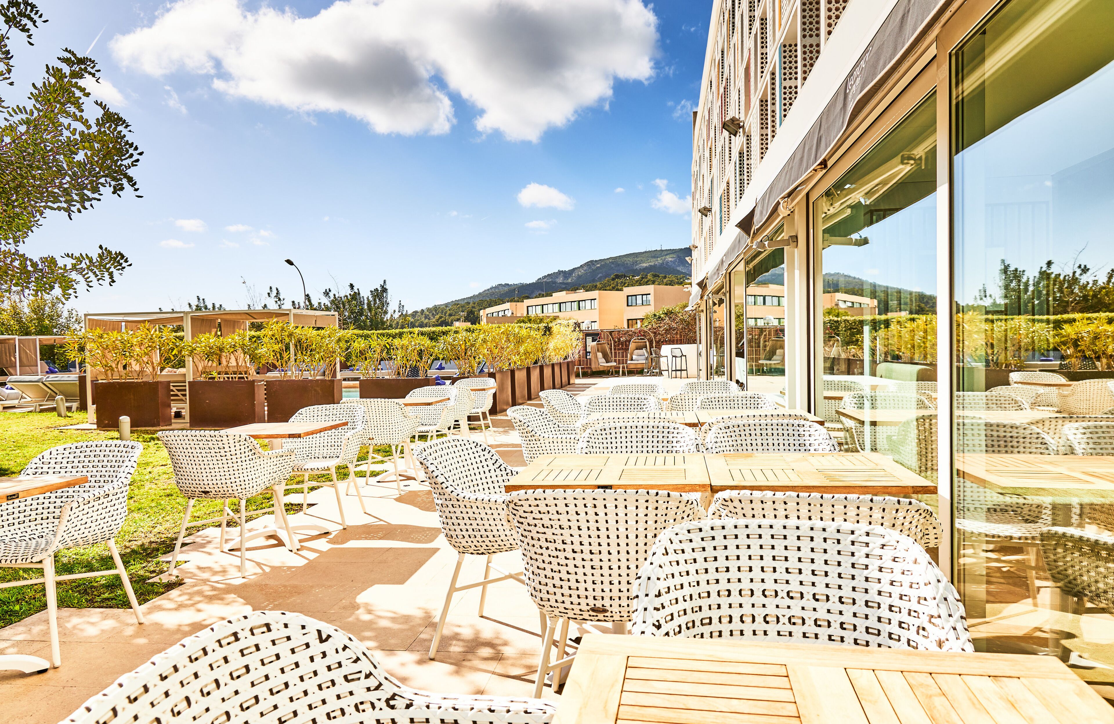 En udendørs terrasse med fletstole og borde, udsigt til omkringliggende bygninger, og en klar himmel.