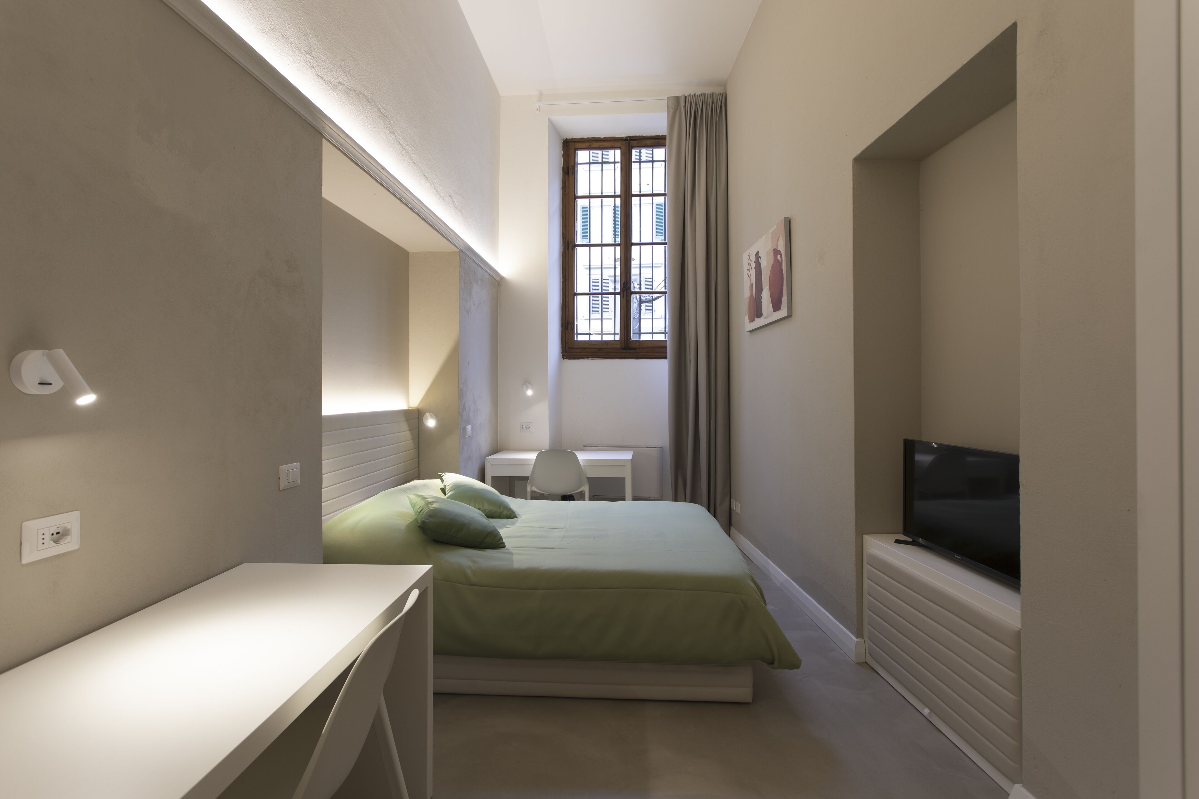 A compact bedroom with a bed, a desk, a chair, a television, and a window with curtains.