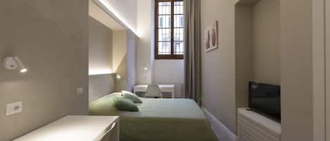 A compact bedroom with a bed, a desk, a chair, a television, and a window with curtains.