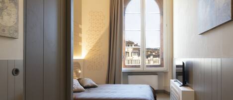 A bedroom with a bed, a radiator, a TV, and a window with a view of buildings.