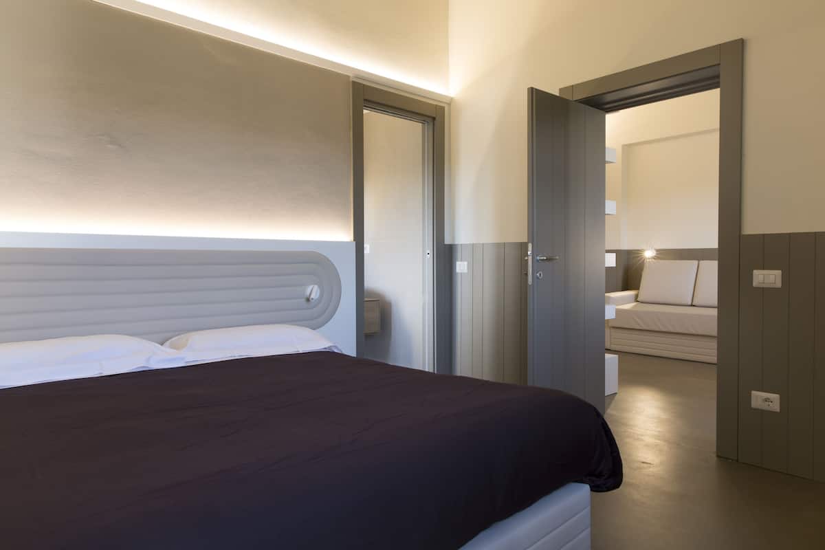 Ein modernes Hotelzimmer mit einem großen Bett, einem Nachttisch und einer Tür zu einem Sitzbereich mit einer Couch.