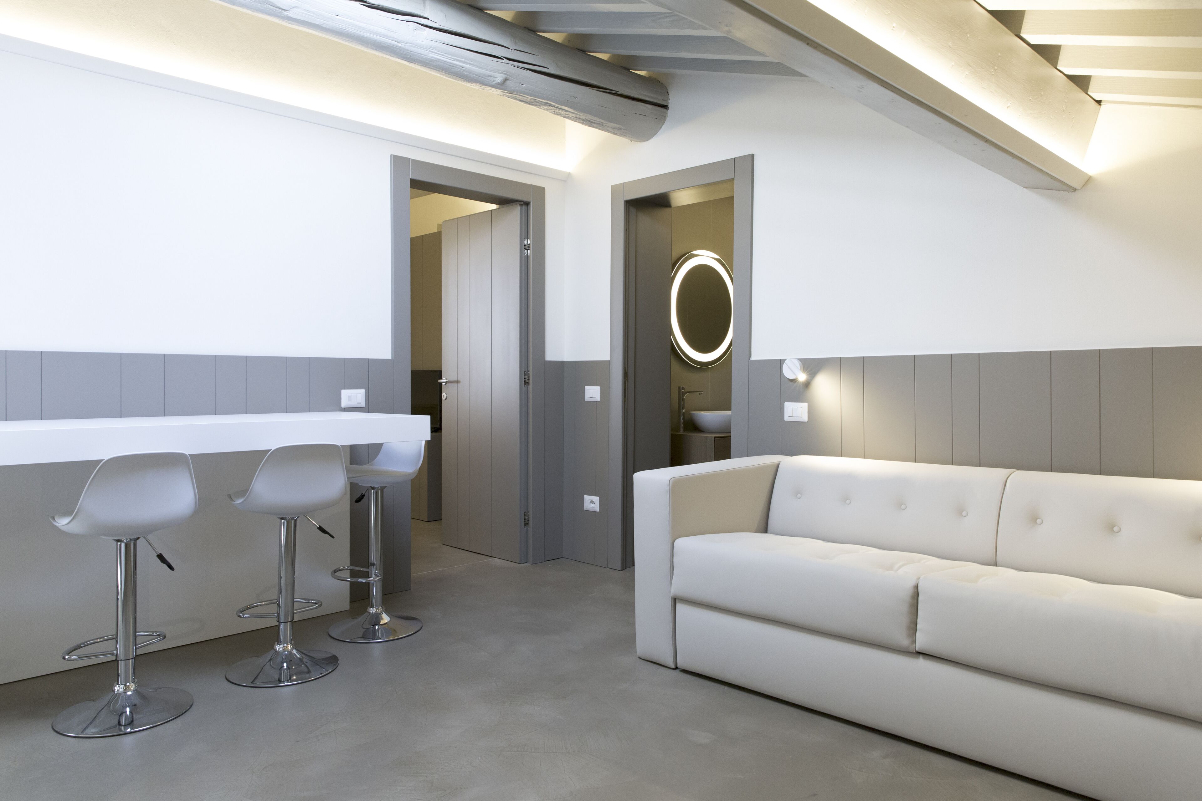 A modern kitchen with a white countertop, three bar stools, a white sofa, and a round mirror above the sink.
