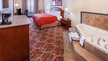 A hotel room with a large bed, a separate sitting area with a chair, a desk with a coffee maker, a bathroom with a bathtub, and a patterned carpet.