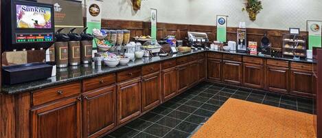 Un espace pour le petit-déjeuner à l’hôtel, avec un buffet, des machines à café et divers récipients contenant de la nourriture.