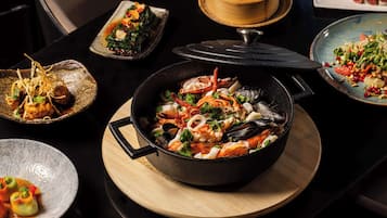 Verschiedene Meeresfrüchtegerichte auf einem Tisch, darunter eine große Pfanne mit Meeresfrüchten, ein Teller mit einem Meeresfrüchtegericht sowie weitere Gerichte mit Gemüse und Meeresfrüchten.