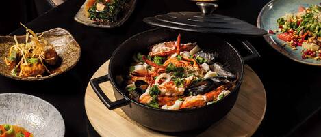 Variedad de platos de mariscos sobre una mesa, incluyendo una paellera grande con mariscos, un plato con preparación de mariscos y otros platos con verduras y mariscos.