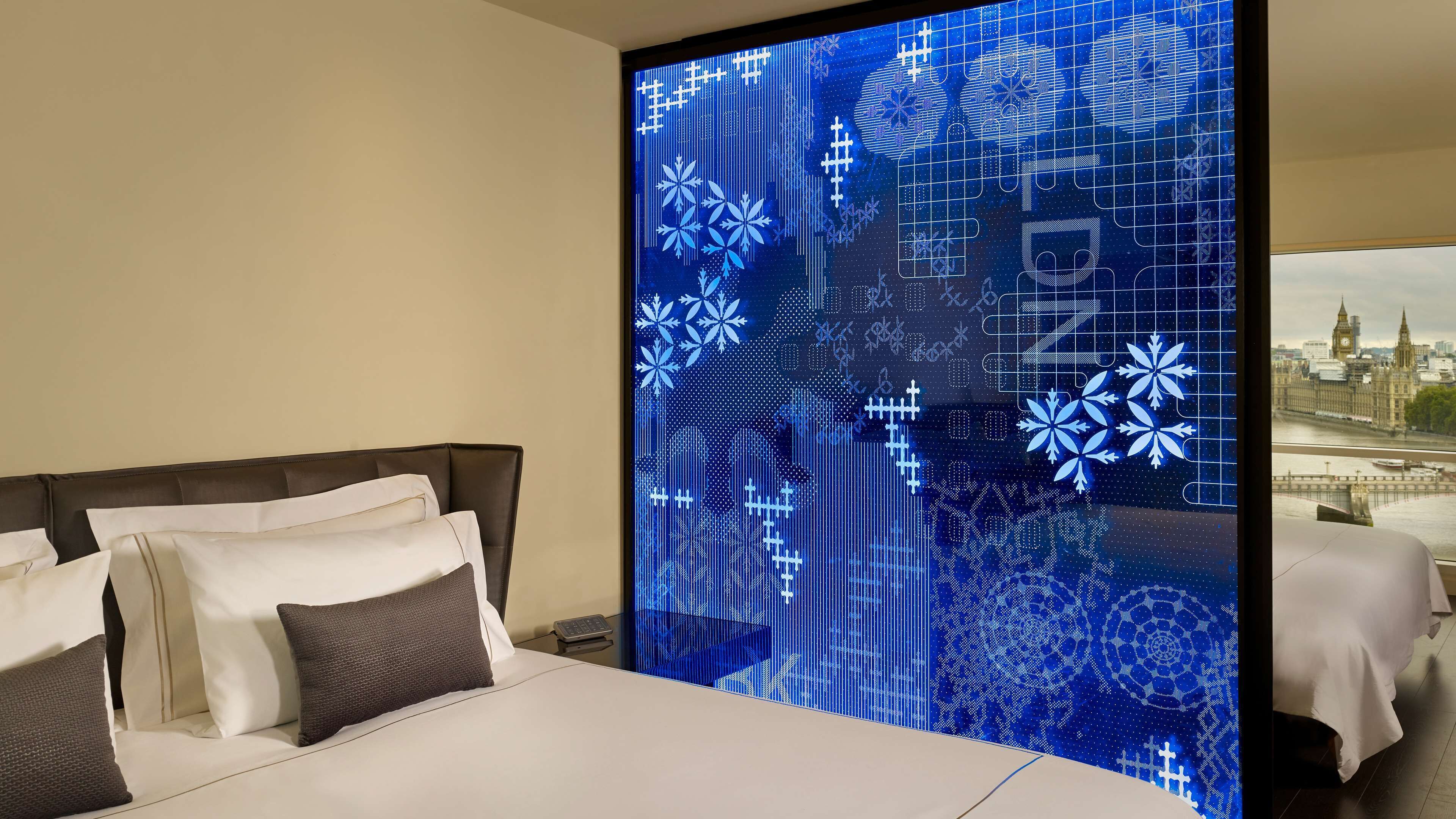 A hotel room with a large bed, a nightstand, and a digital display wall with a cityscape view.