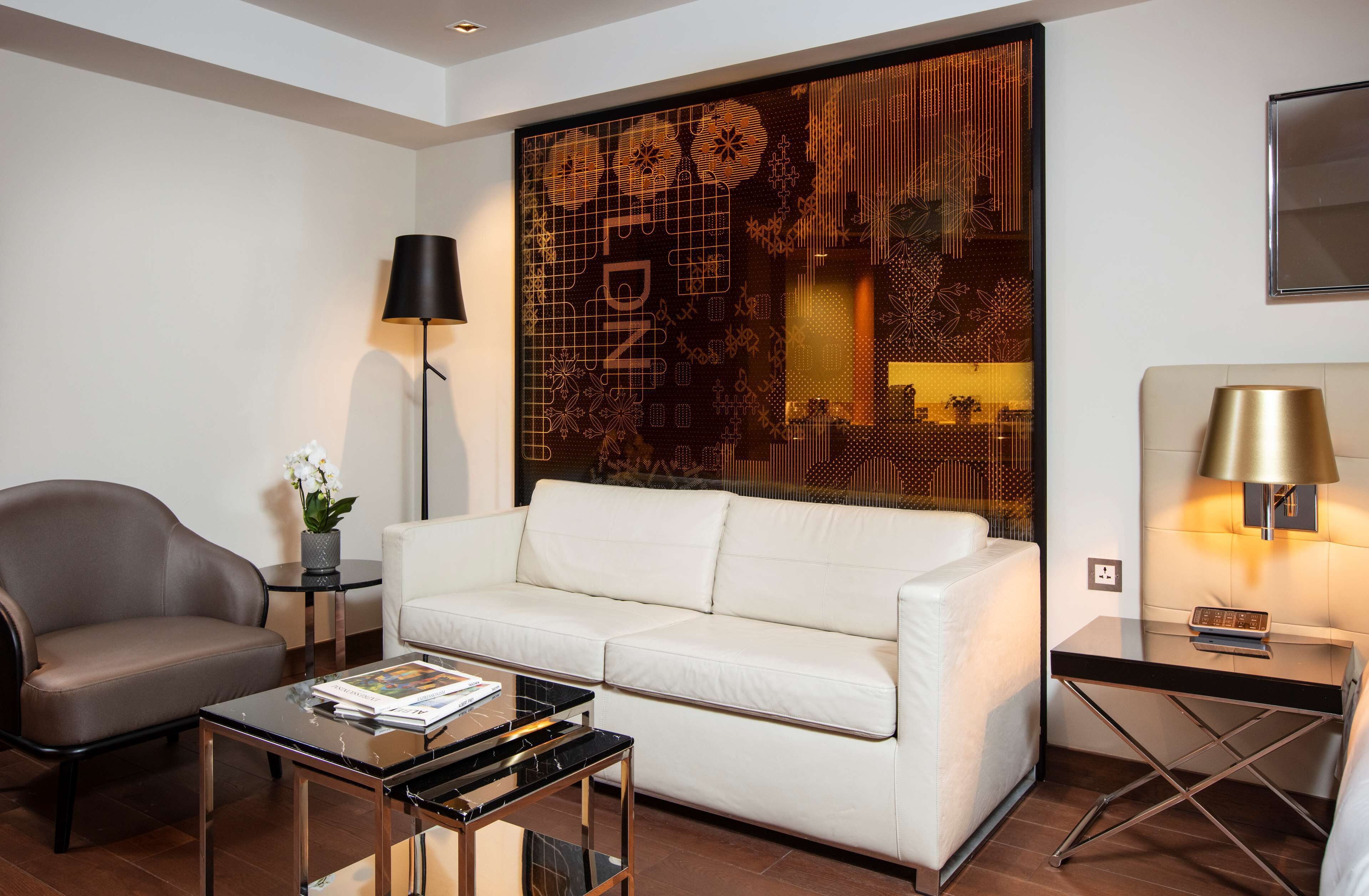 A modern living room with a white sofa, a brown armchair, a glass coffee table, and a decorative wall panel.
