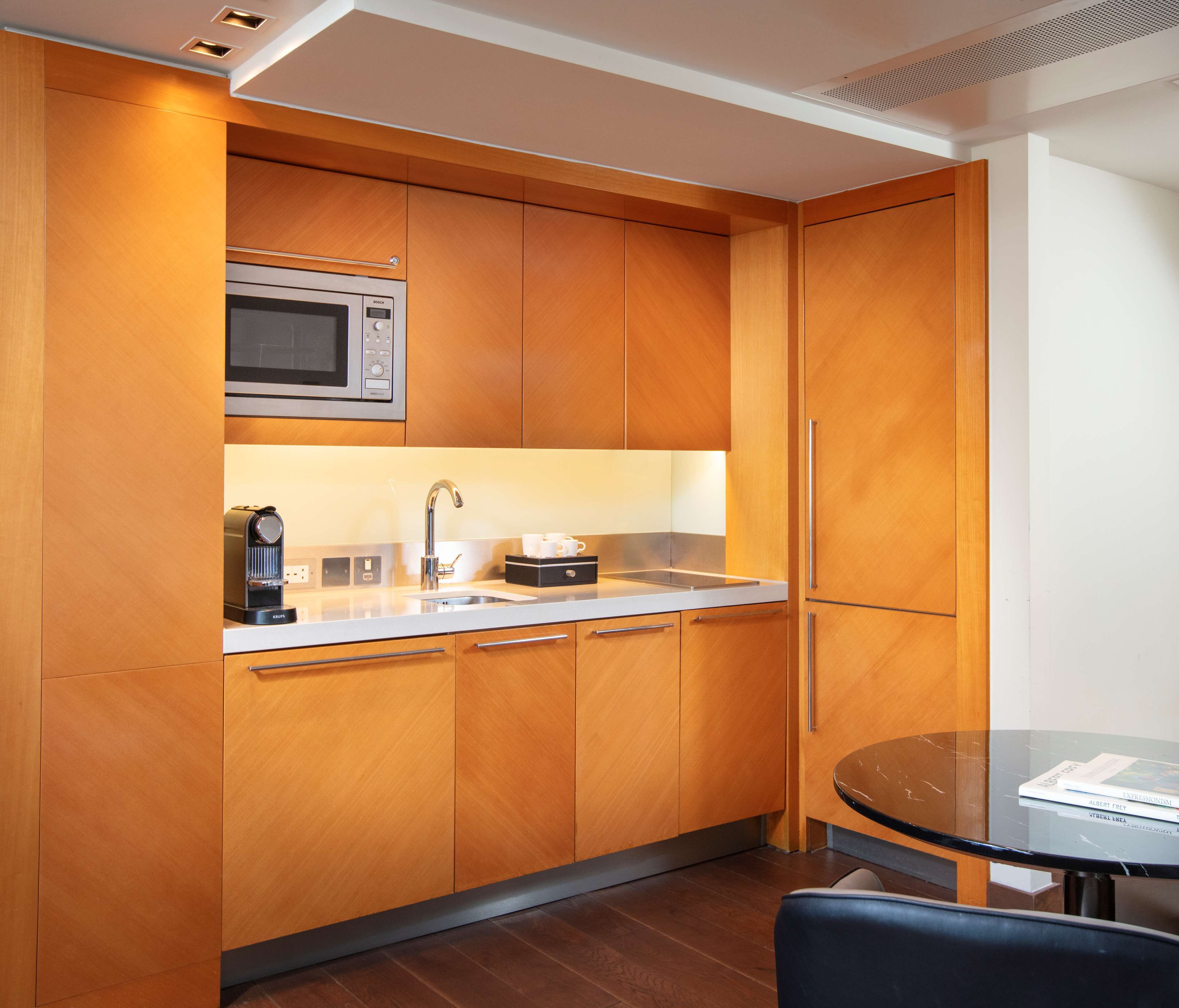 A modern kitchen with wooden cabinets, a stainless steel microwave, a coffee maker, and a glass table with a chair.