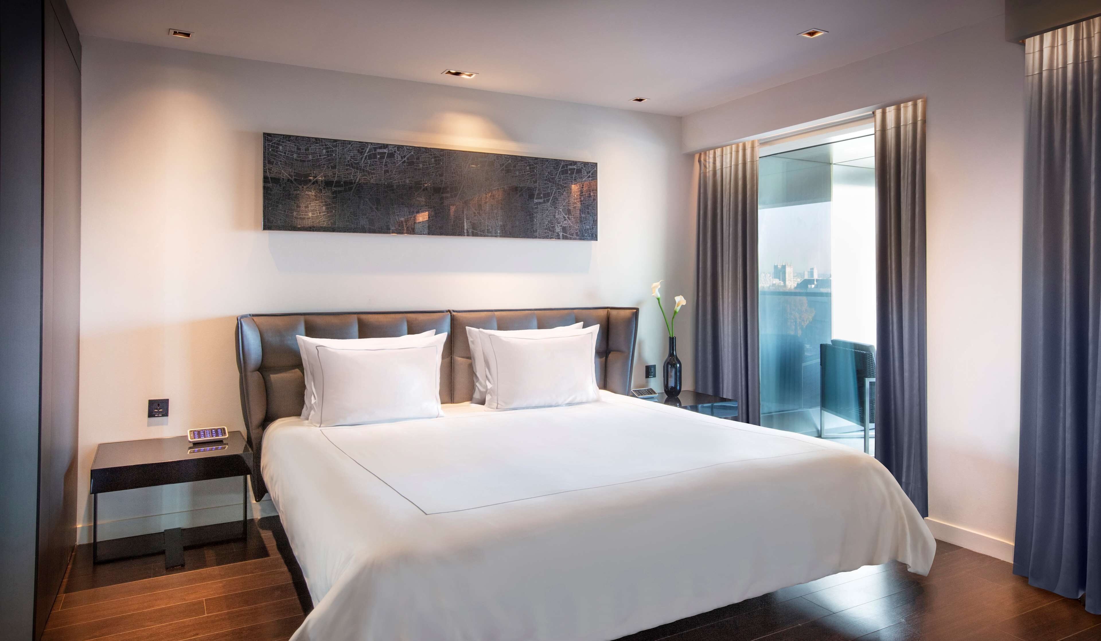 A modern hotel room with a large bed, a nightstand, a vase with flowers, and a view of the city through the window.