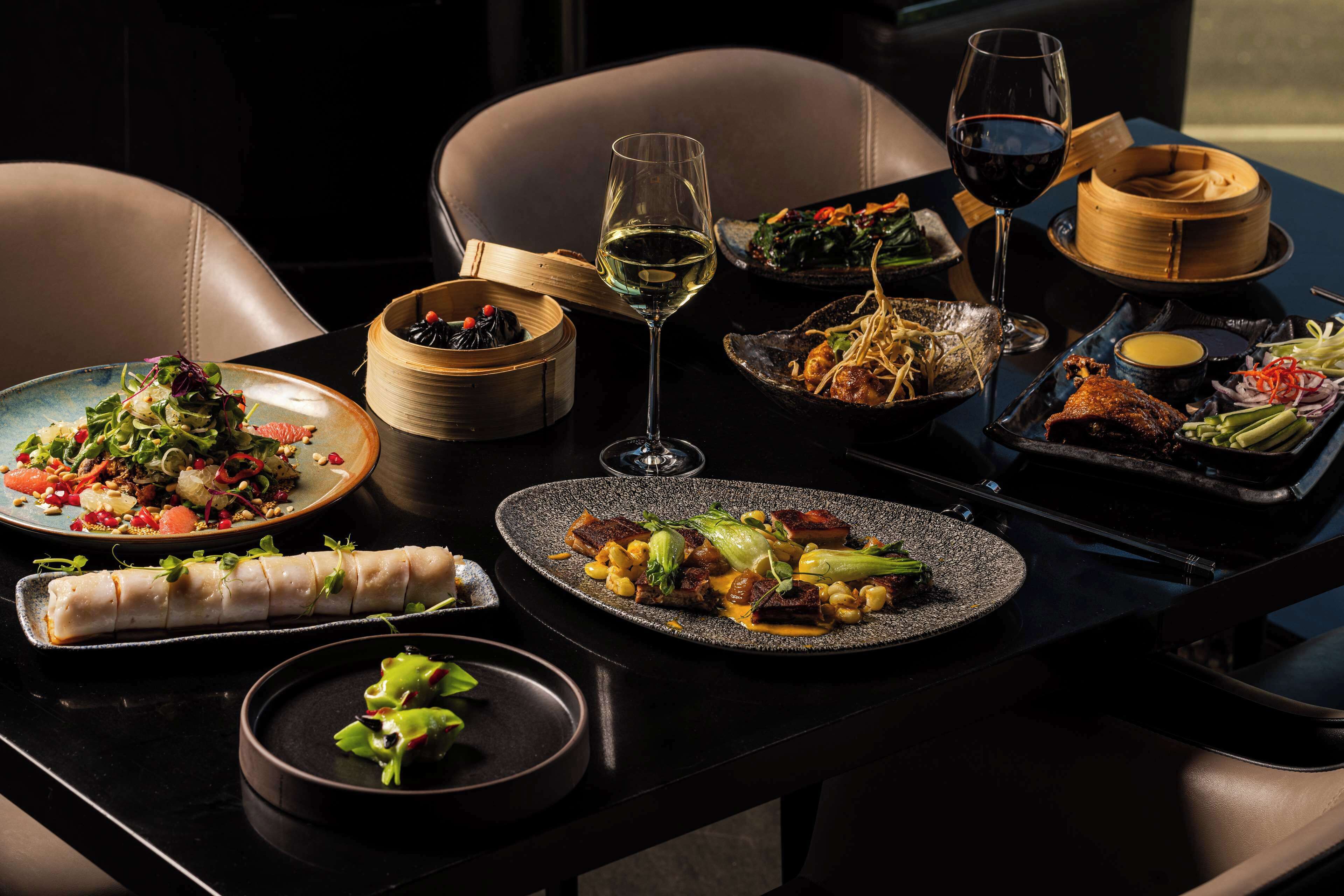 A table set with various dishes, including a rolled appetizer, a salad, and a plate with meat and vegetables, along with two glasses of wine.
