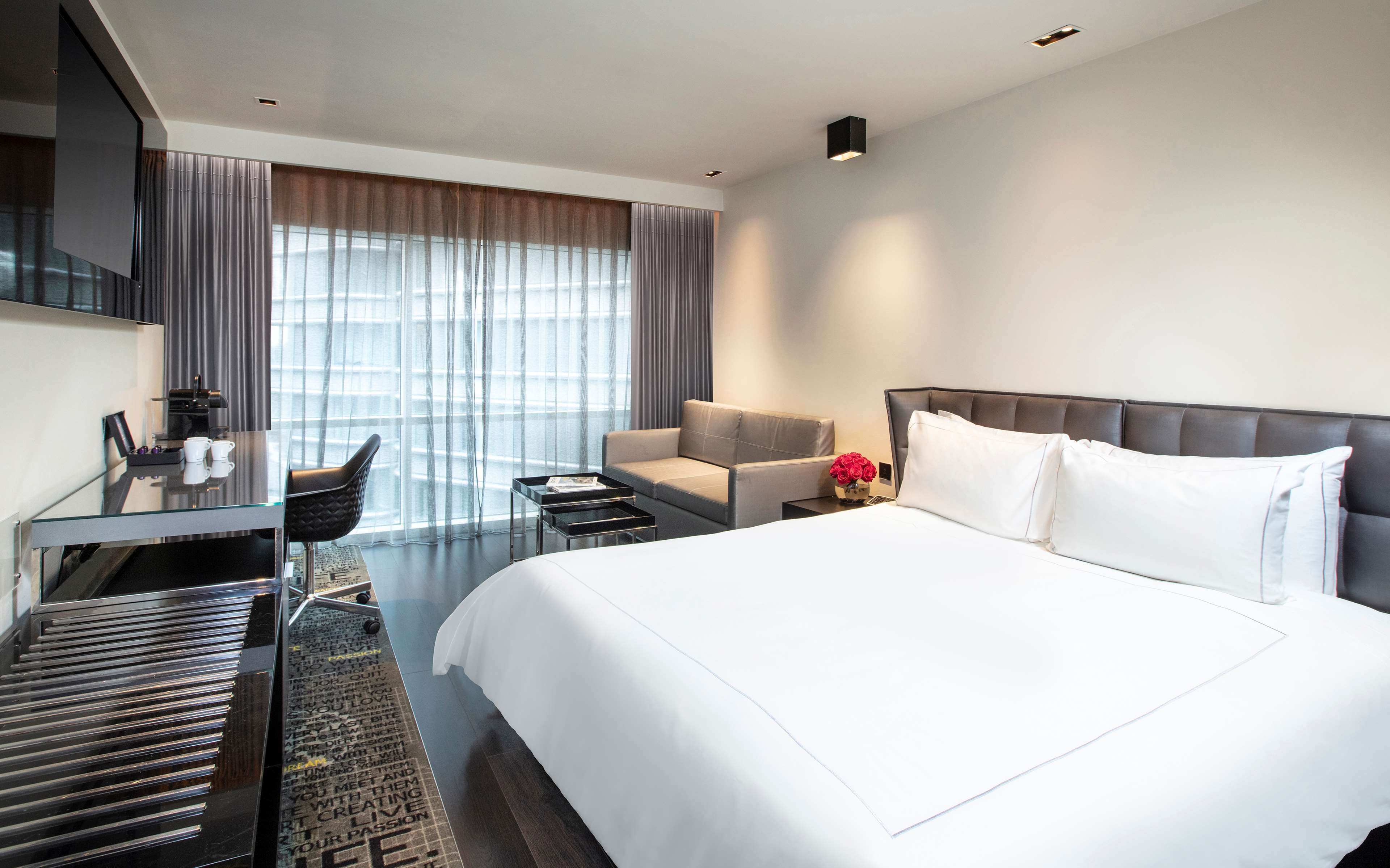 A modern hotel room with a large bed, a sofa, a desk with a chair, and a TV mounted on the wall.