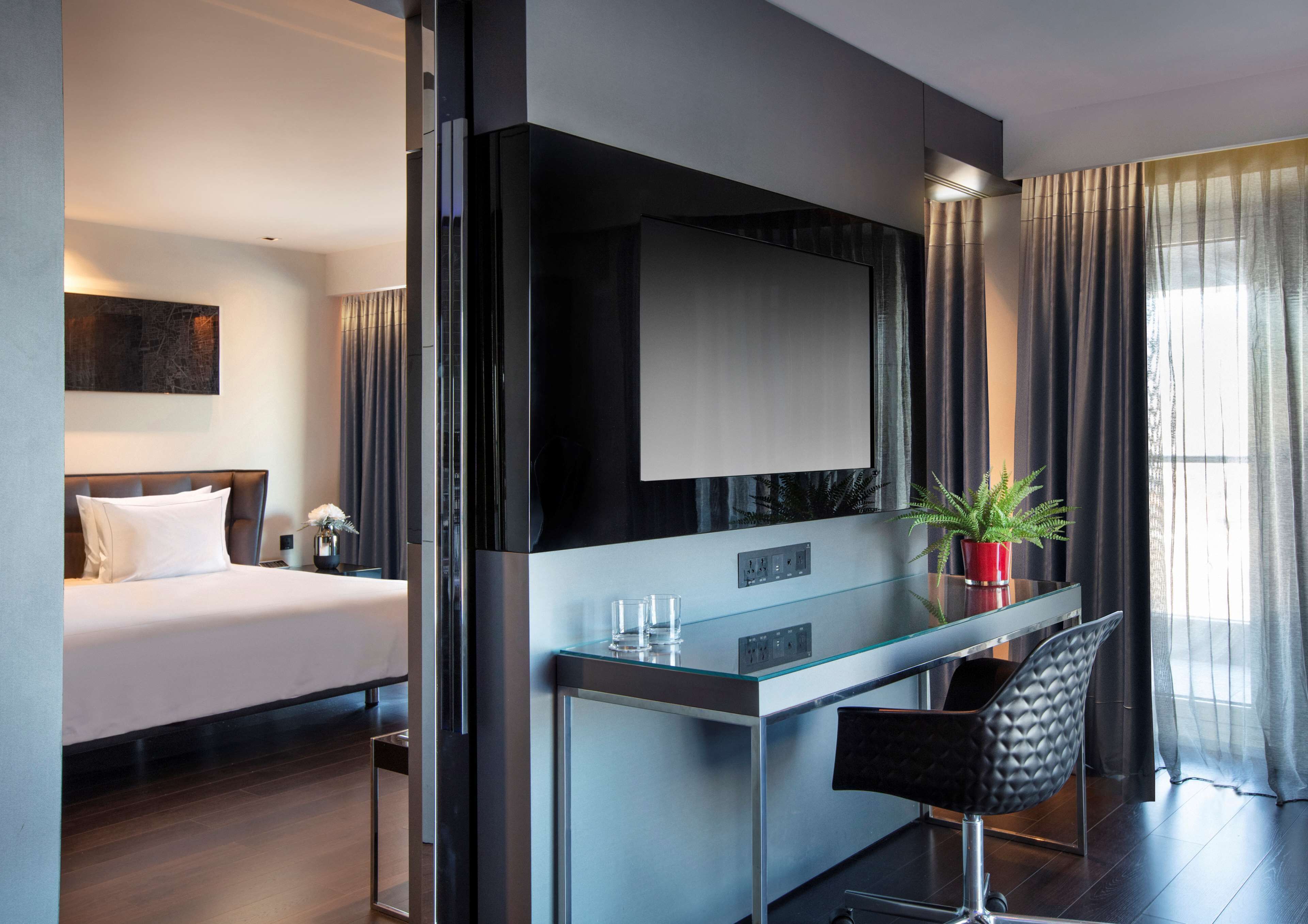 A modern hotel room with a bed, a glass desk, a chair, a TV, and a potted plant.