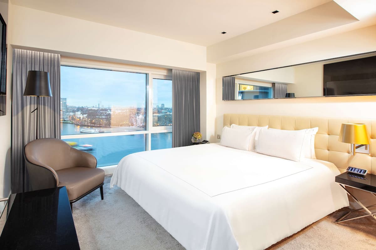 Habitación de hotel con una cama grande, una silla, un escritorio y vistas de un paisaje urbano a través de amplias ventanas.