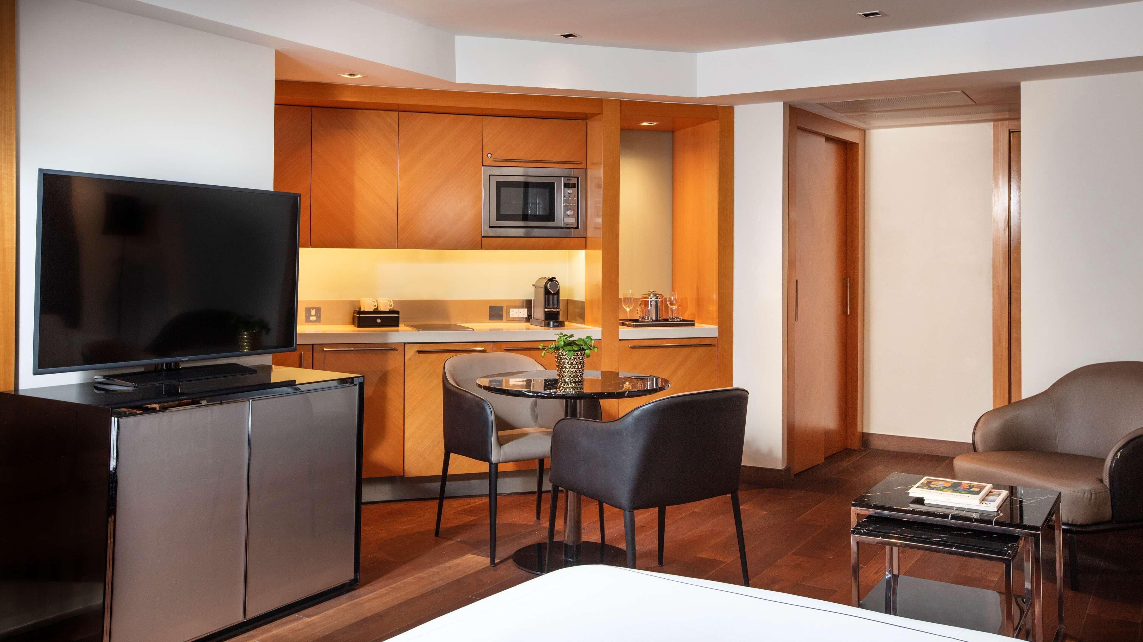 A compact hotel room with a kitchenette, a flat-screen TV, a dining area with a table and chairs, and a seating area with a sofa.