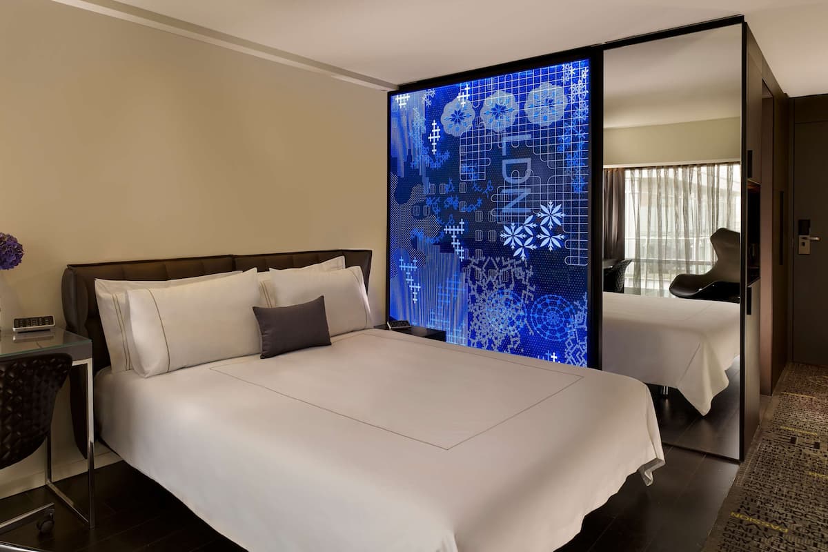 Habitación de hotel con una cama grande, un escritorio con un jarrón de flores y una pantalla digital en la pared.
