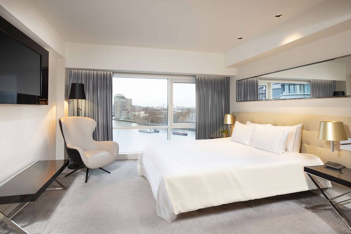 Habitación de hotel moderna con una cama grande, un escritorio, una silla, un televisor y un ventanal con vistas a la ciudad.