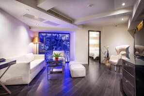 Una habitación de hotel moderna con un sofá blanco, una mesa de centro de cristal, una pequeña planta en maceta, una pared decorativa con una pantalla digital y un espejo grande.
