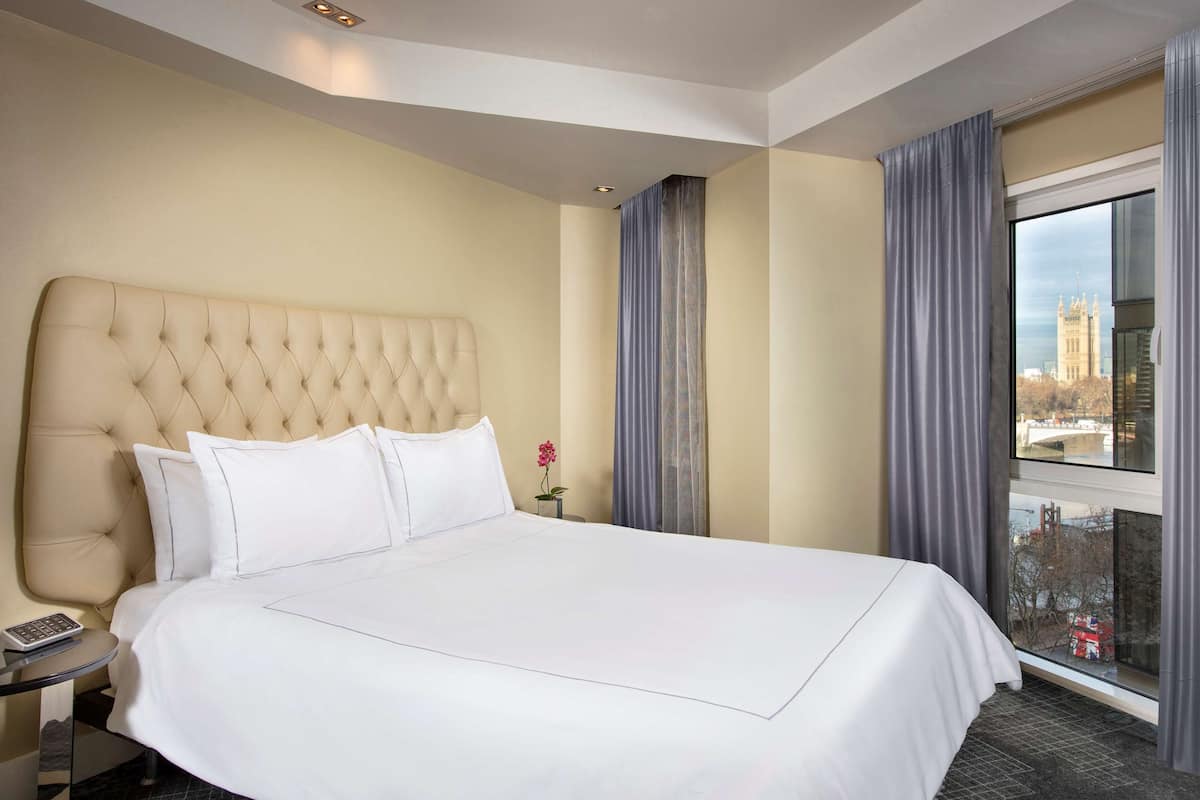 Habitación de hotel con una cama grande, una mesita de noche, una ventana con cortinas y vistas a un paisaje urbano.