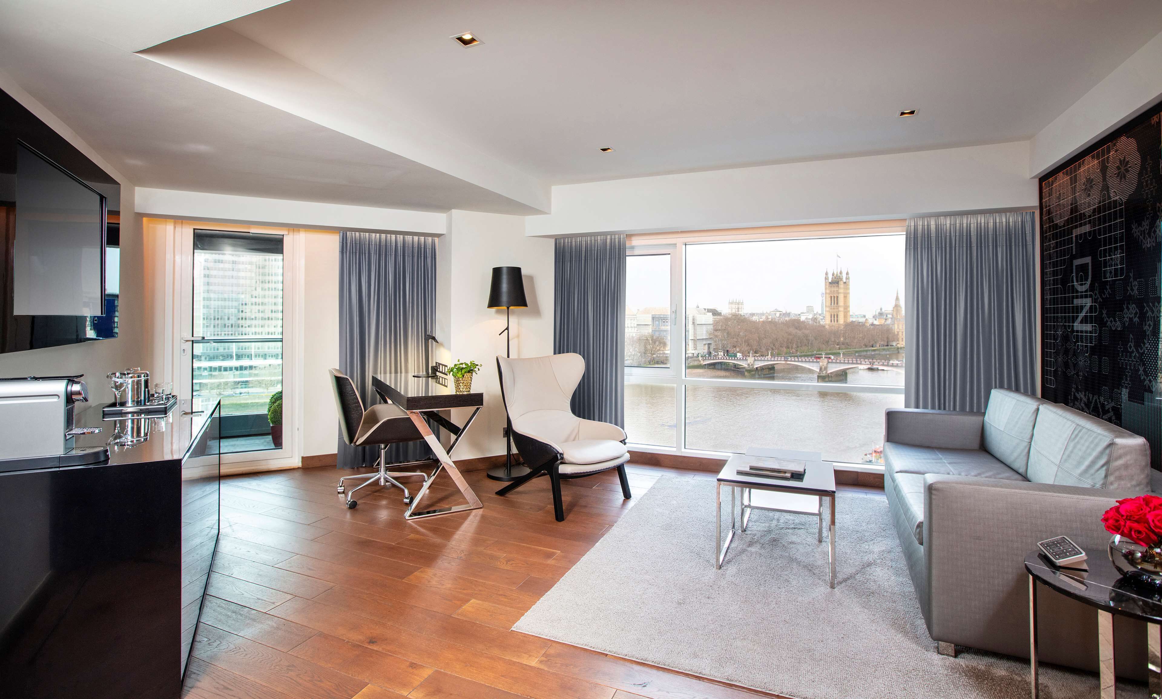 Ein modernes Wohnzimmer mit Sofa, Esstisch mit Stühlen, Küchenbereich und großen Fenstern mit Blick auf eine Stadtlandschaft.