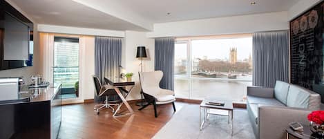 A modern living room with a sofa, a dining table with chairs, a kitchen area, and large windows offering a view of a cityscape.