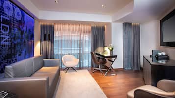 A modern hotel room with a large digital display, a sofa, a desk with a lamp, and a chair.