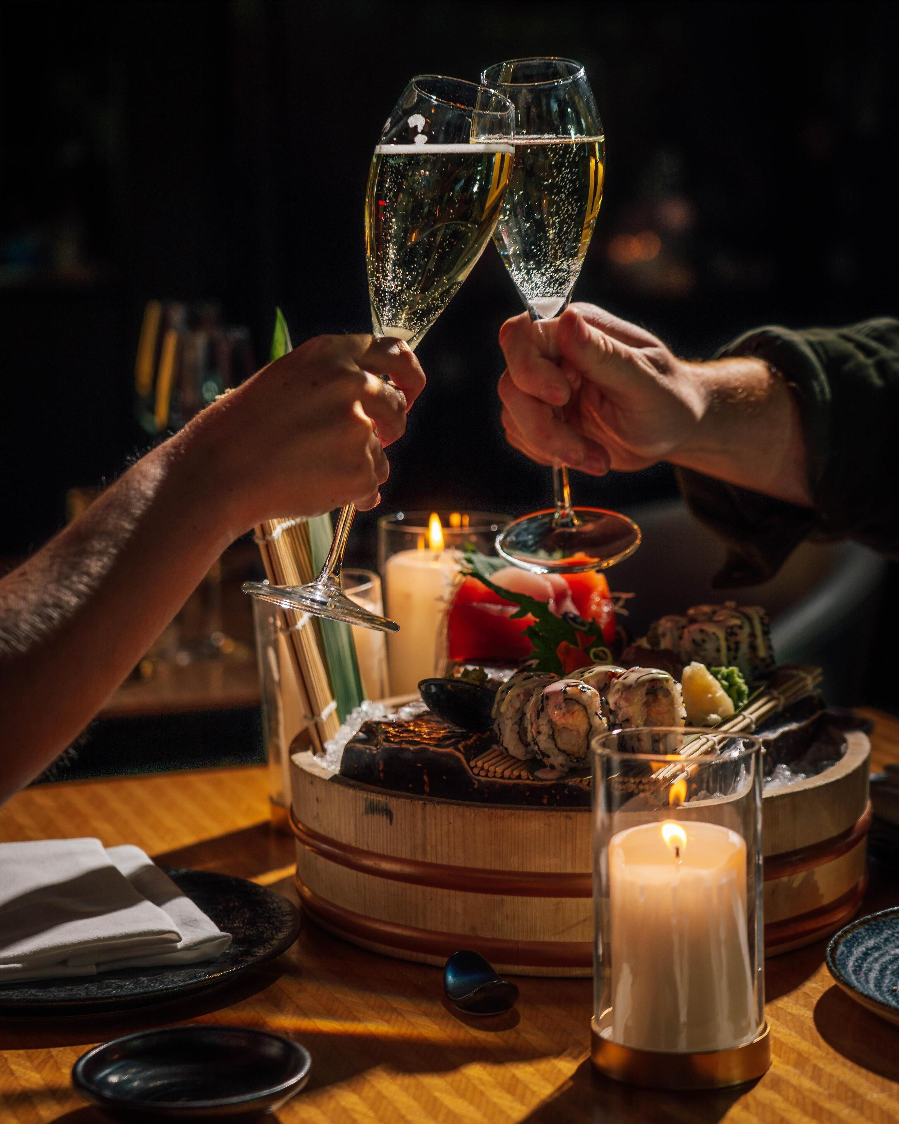 A wooden table with sushi, a candle, and two glasses of champagne being toasted.