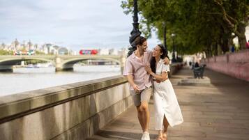 A couple walking along a riverside promenade with a bridge and cityscape in the background.