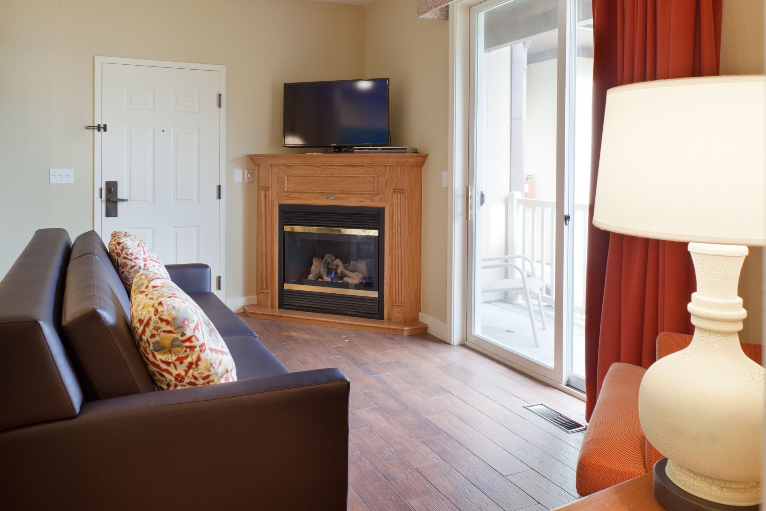 A living room with a fireplace, a television on a stand, a sofa with patterned cushions, and a table with a lamp.