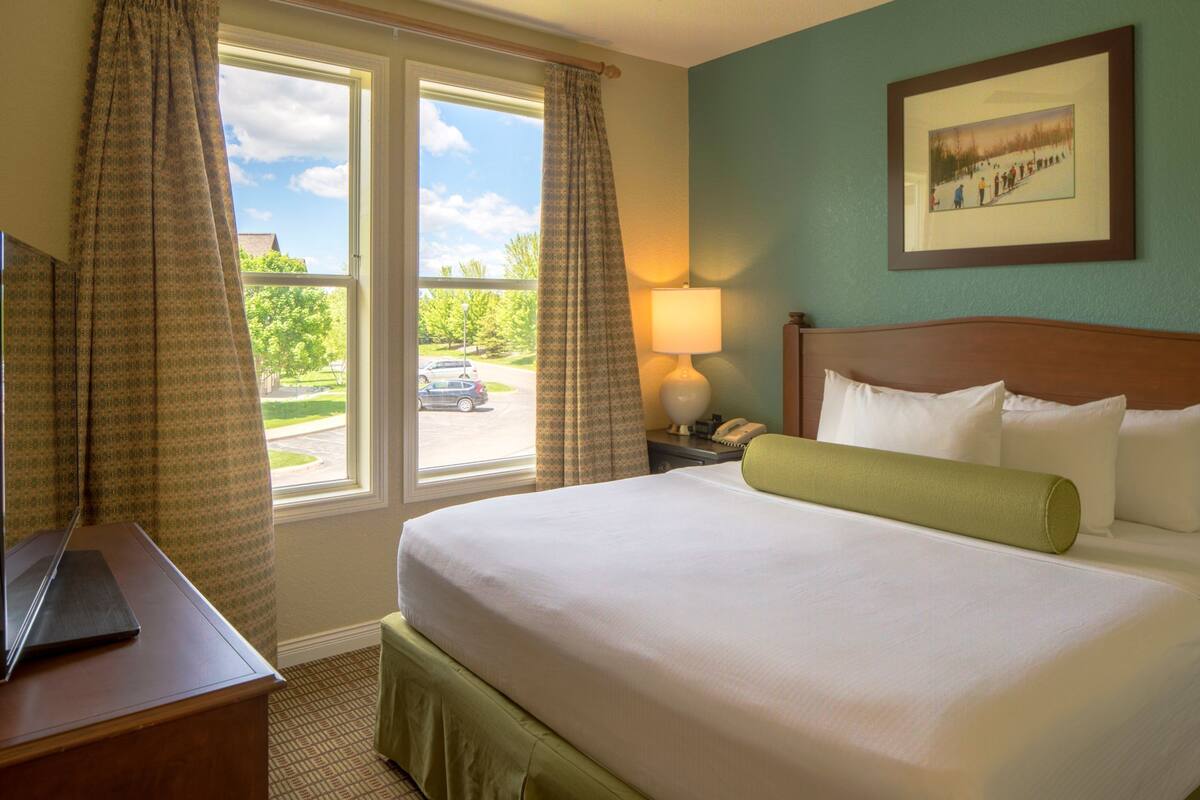 A hotel room with a bed, a desk with a laptop, a lamp, and a window with curtains.