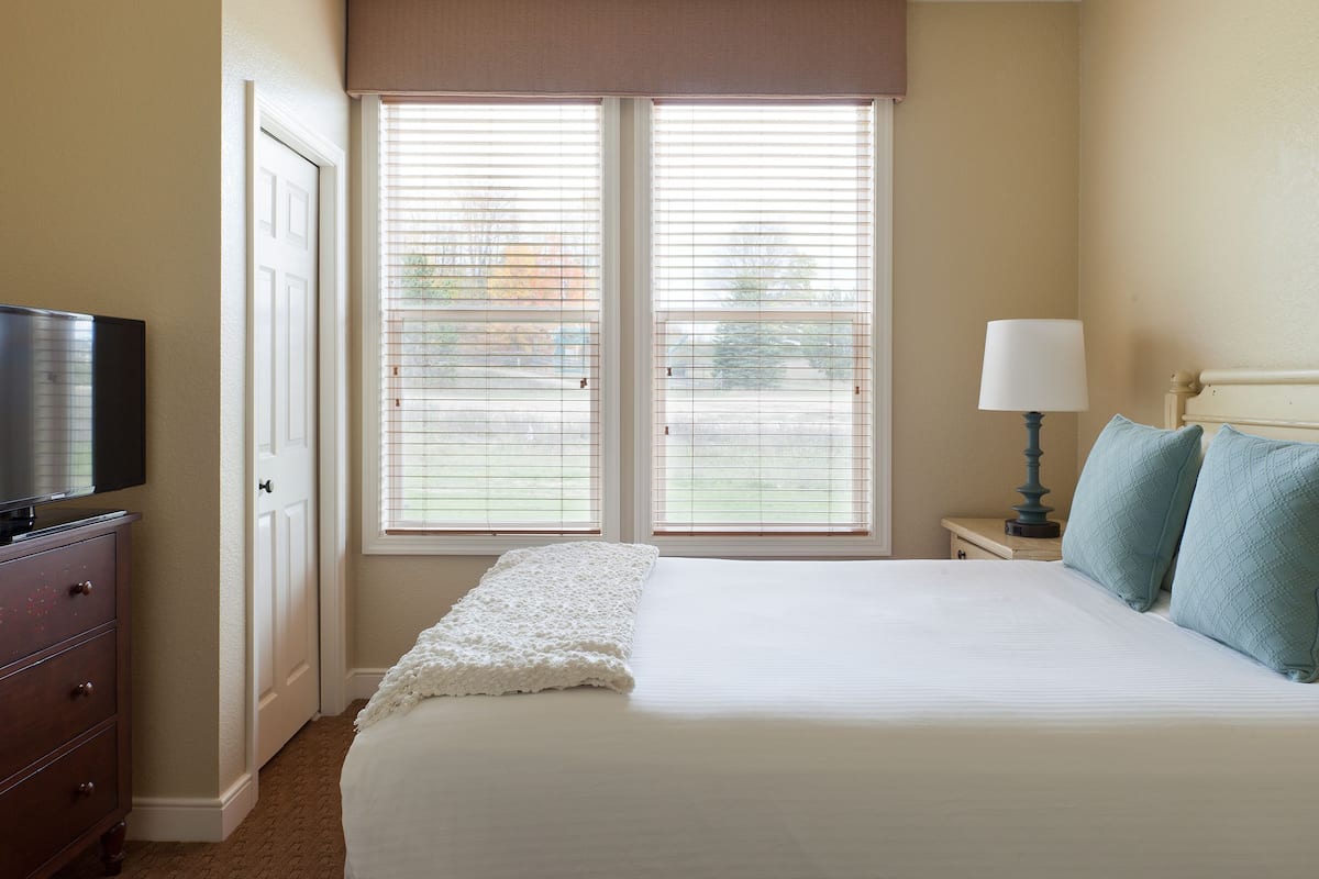 A neatly made bed with a white comforter and two pillows, a bedside lamp, and a television on a dresser in a room with beige walls and a window with blinds.