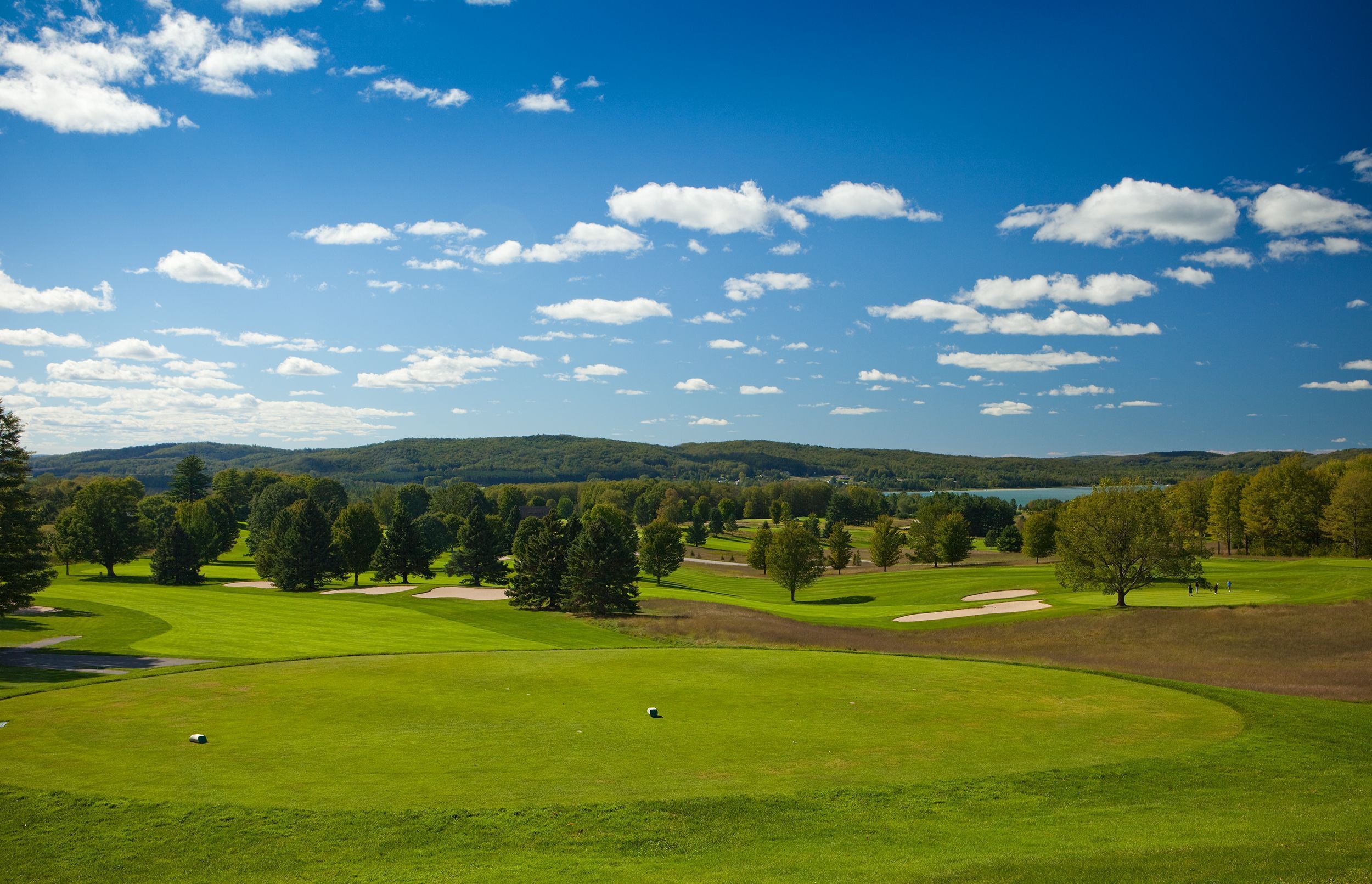 Ein Golfplatz mit blauem Himmel, grünem Rasen, Bäumen und einem See in der Ferne.