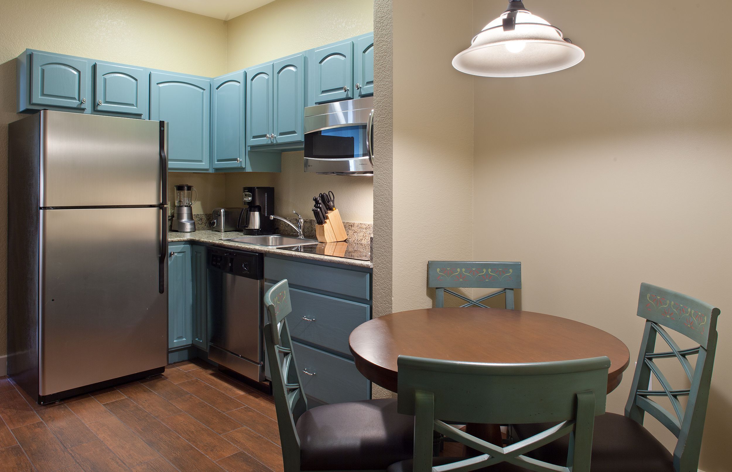 A compact kitchen with teal cabinets, a stainless steel refrigerator, a microwave, and a dining area with a round table and two chairs.