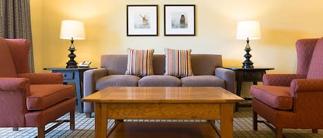 A cozy room with a sofa, two armchairs, a wooden coffee table, and two lamps. There are framed pictures on the wall.