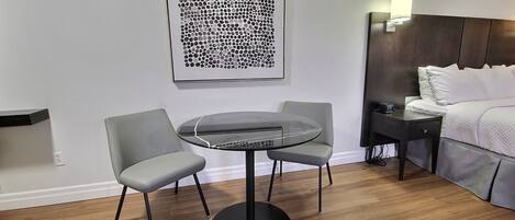 A modern hotel room with a round table, two chairs, a bed with white linens, a nightstand, and a wall-mounted artwork.
