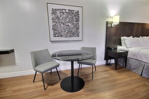 Una habitación de hotel moderna con una mesa redonda, dos sillas, una cama con ropa blanca, una mesita de noche y una obra de arte colgada en la pared.