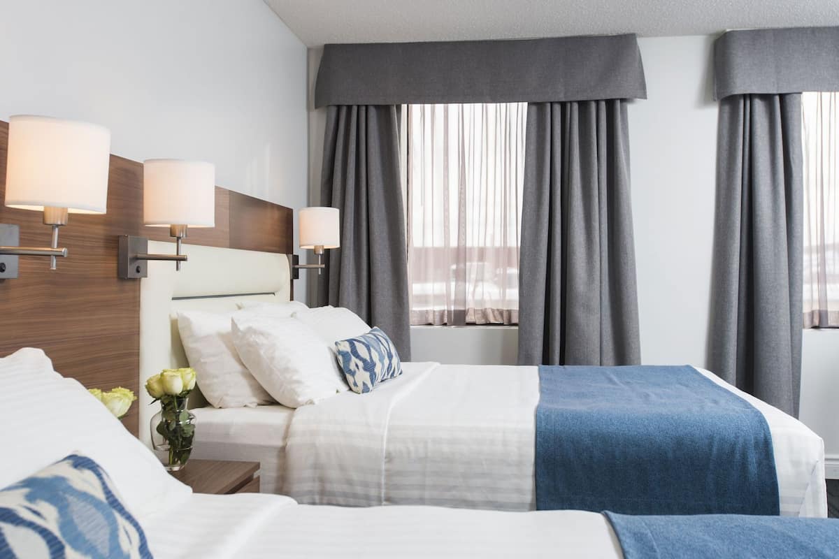 Habitación de hotel con dos camas, cabecero de madera, lámparas fijadas a la pared y un ventanal con cortinas grises.