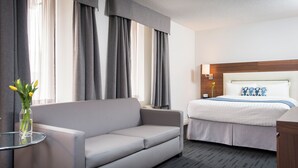 Ein Hotelzimmer mit grauem Sofa, einem Bett mit weißem Kopfteil und einem kleinen Tisch mit Blumen.