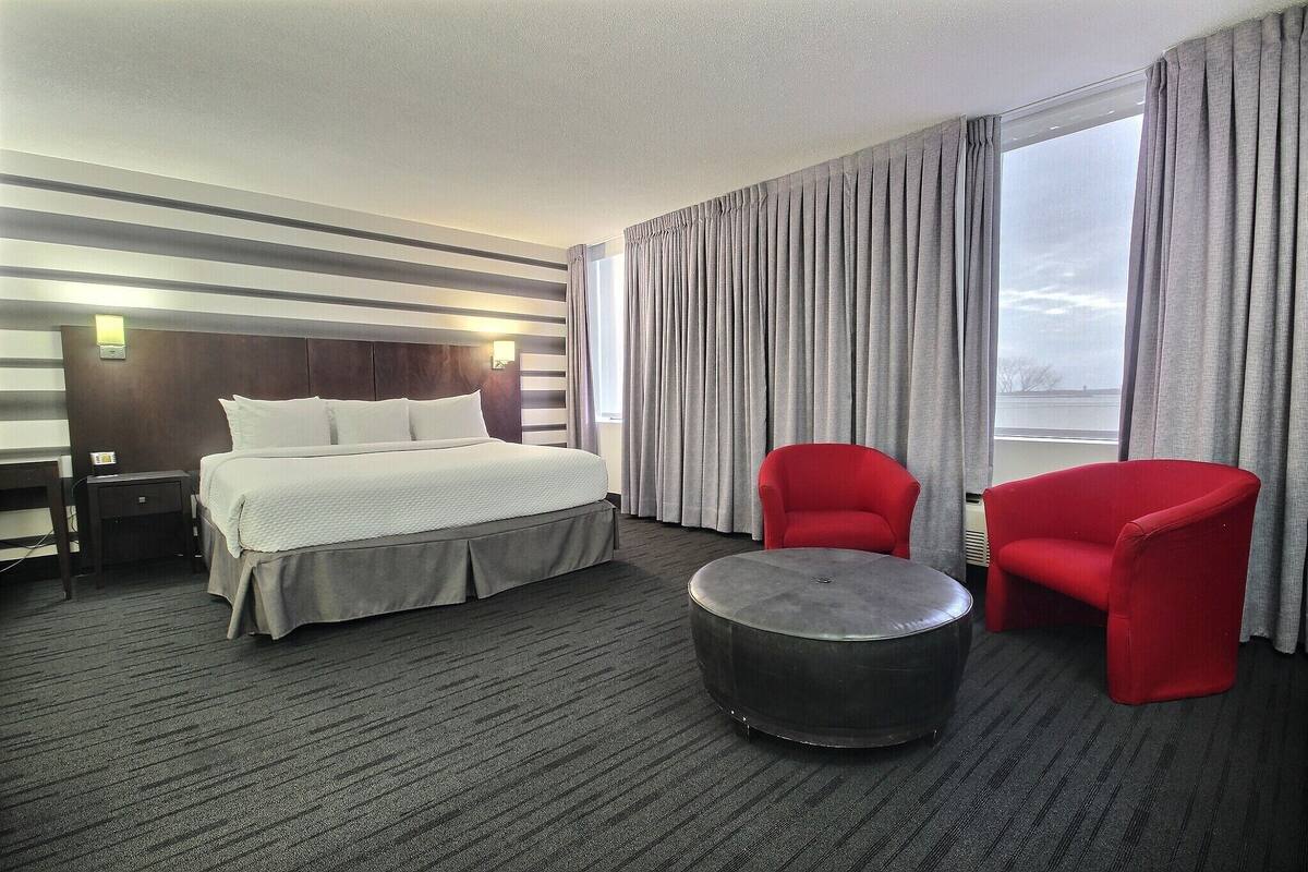 Habitación de hotel con una cama, dos sillas rojas, una mesa redonda y vistas al exterior a través de amplias ventanas.