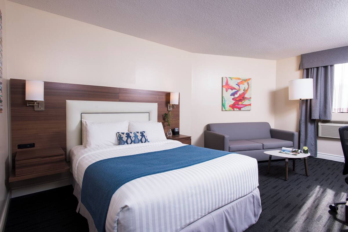 Una habitación de hotel con una cama grande, un sillón gris, una mesita y un cuadro en la pared.