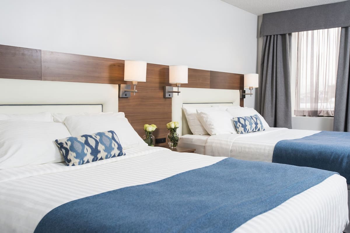 Habitación de hotel con dos camas, cabecera de madera, lámparas de pared y ventana con cortinas.