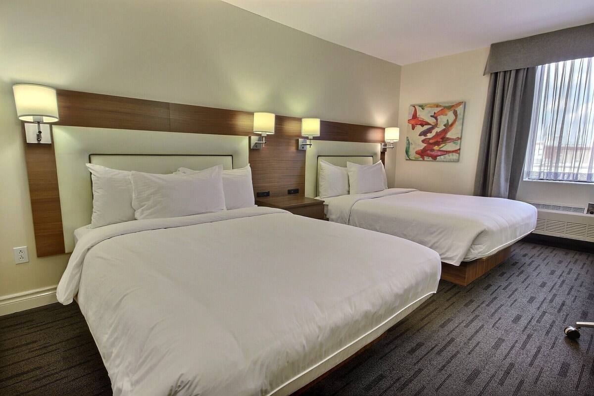 Habitación de hotel con dos camas, cabecero de madera, lámparas de pared y un ventanal con cortinas.