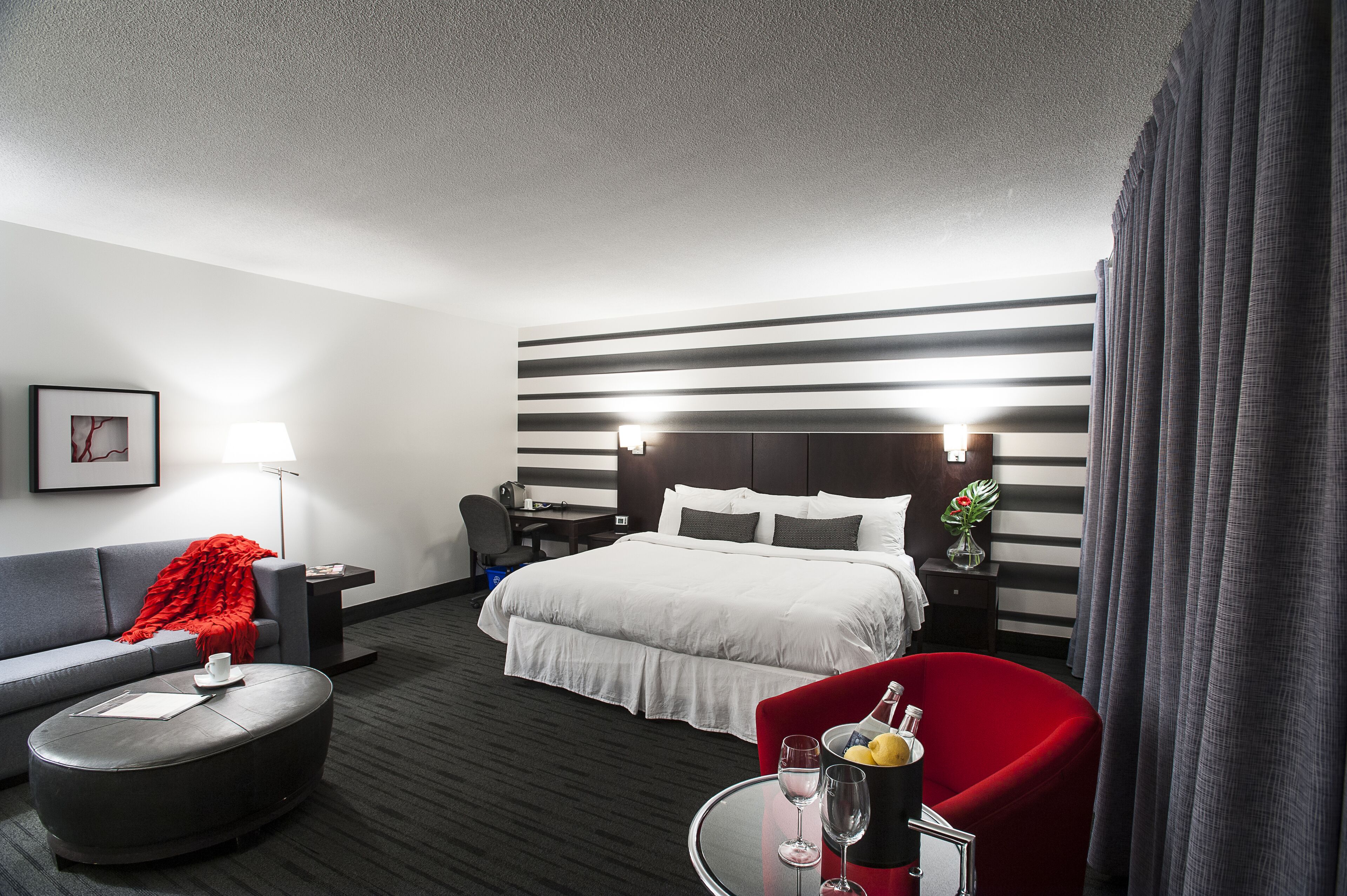 A hotel room with a large bed, a sofa, a red armchair, a small table with a bottle and glasses, and a desk with a lamp.